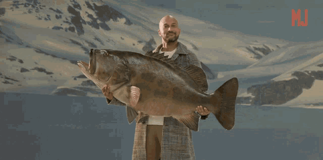 a man is holding a large fish in front of a mountain with the letter m on it