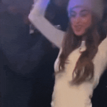 a woman in a white dress and hat is dancing in a dark room with her arms in the air .