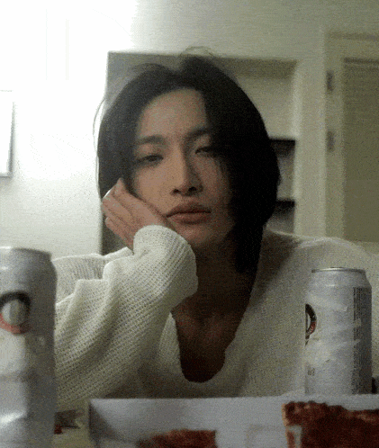 a woman in a white sweater sits at a table with two cans of beer and a pizza