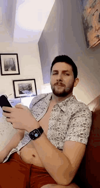 a man without a shirt is sitting on a couch looking at his cell phone .