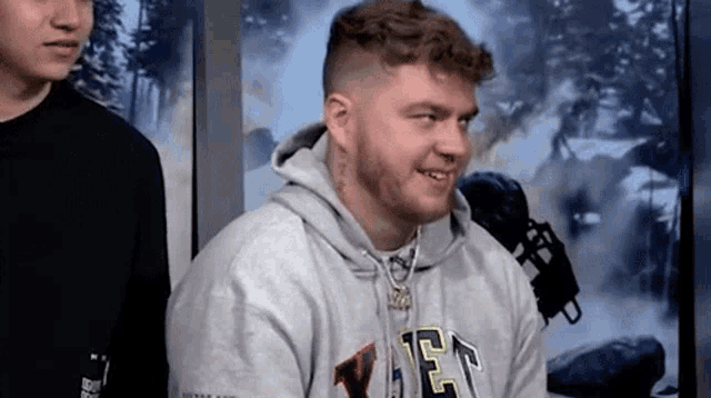a man with a beard is wearing a gray hoodie with the letter e on it .