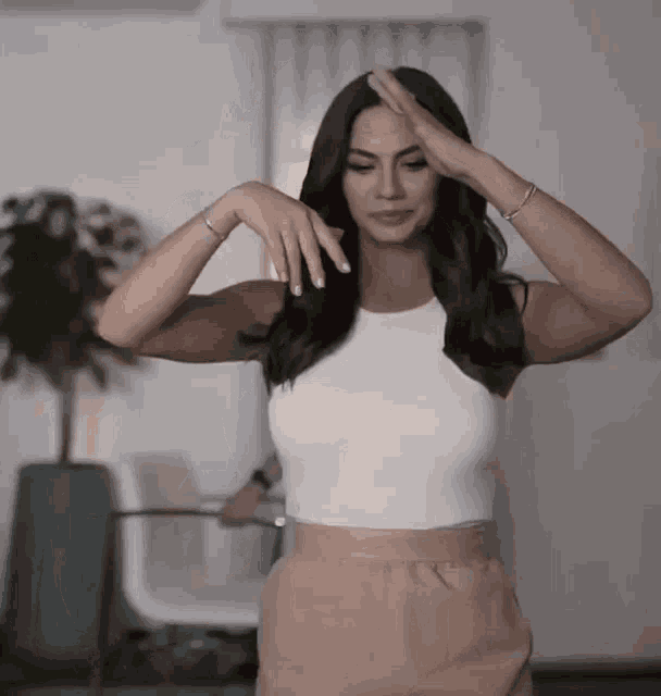 a woman wearing a white tank top and pink pants holds her hand to her head