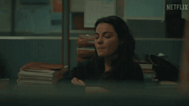 a woman sits at a desk with her arms crossed and a netflix logo visible in the background