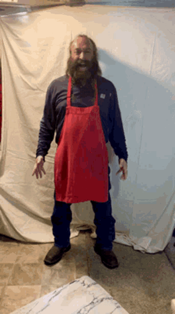 a man with a beard wearing a red apron stands in front of a white cloth