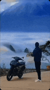 a man is standing next to a motorcycle in front of a mountain covered in clouds .