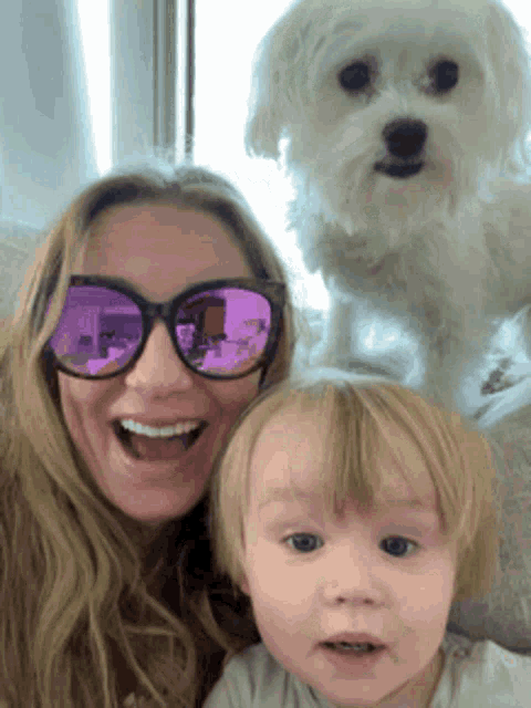 a woman wearing purple sunglasses holds a baby and a dog
