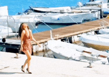 a woman in a red dress is walking on a pier with boats in the background