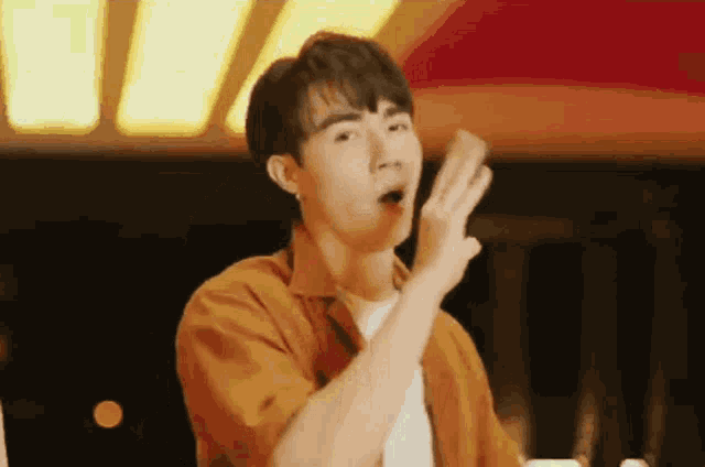 a young man in an orange shirt is making a funny face while holding a piece of paper in his hand .