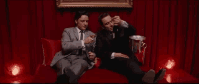 two men in suits are sitting on a red couch drinking champagne and toasting .