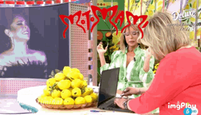 two women are sitting at a table with a laptop and a basket of lemons