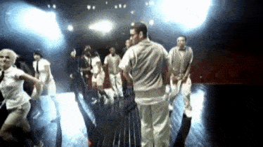 a group of people are dancing on a stage while a man stands in the middle .