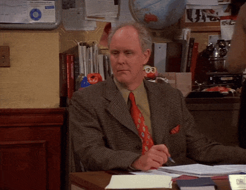 a man in a suit and tie sits at a desk in front of a bulletin board that says " new york times "