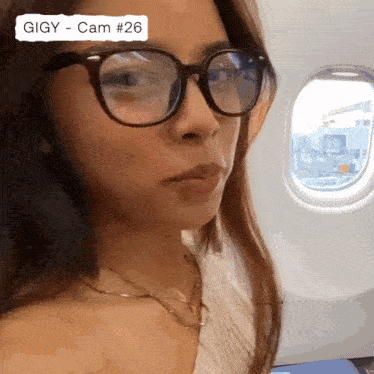 a woman wearing glasses and a necklace is sitting on an airplane with gigiy cam # 26 written on the bottom