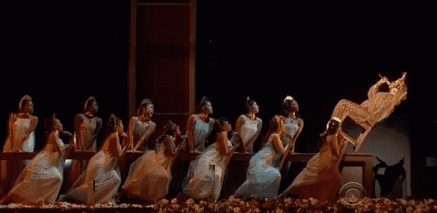 a group of women in white dresses are dancing on a stage with a man in a chair behind them