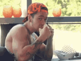 a shirtless man wearing a hat is sitting at a table eating a sandwich .