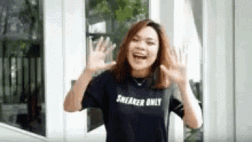 a woman wearing a black sneaker only t-shirt is standing in front of a window and waving .