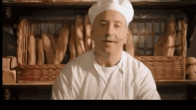 a man with a mustache wearing a chef 's hat stands in front of a shelf of bread