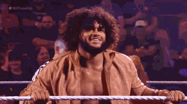 a man in a brown jacket is standing in a wrestling ring with # nxtlevelup written on the bottom