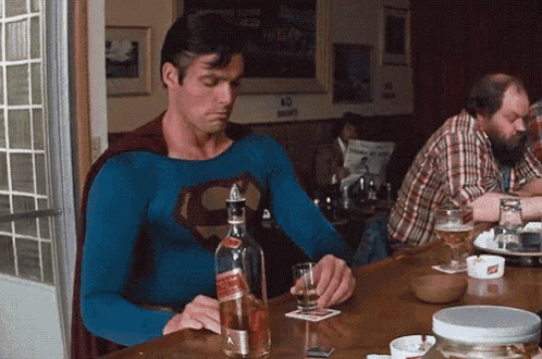 a man dressed as superman is sitting at a bar holding a bottle of alcohol