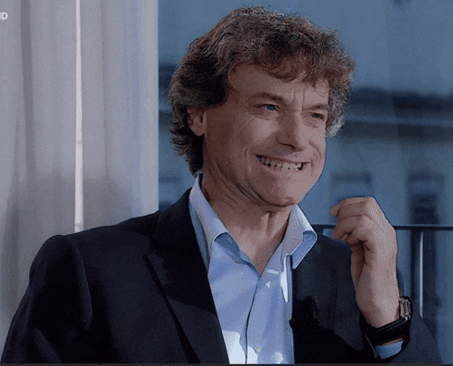 a man in a suit and blue shirt smiles in front of a window with the letters id on the bottom right