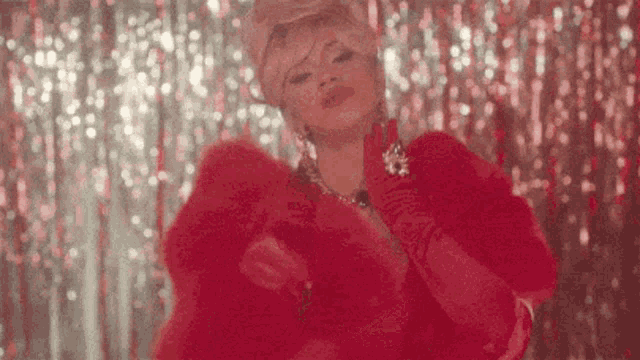 a drag queen in a red fur coat and gloves is standing in front of a red curtain .