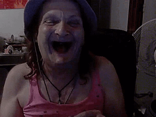 a woman wearing a pink tank top and a blue hat is laughing with her mouth open