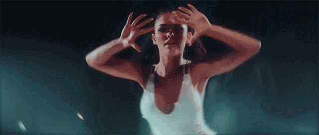 a woman in a white tank top is dancing in a dark room with her hands in the air .