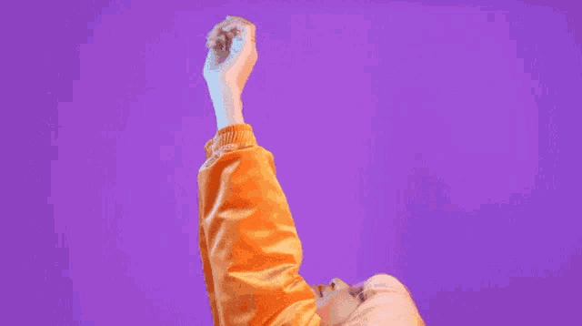 a woman in an orange jacket is reaching up with her hands in the air against a purple background .