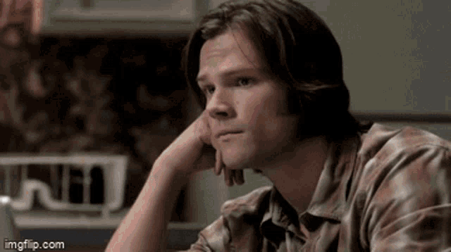 a man with long hair is sitting at a table with his hand on his chin .