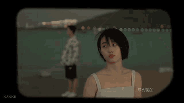 a woman in a white tank top stands in front of a man on a beach with nanke written on the bottom right