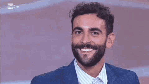 a man with a beard is smiling and wearing a blue suit and white striped shirt .