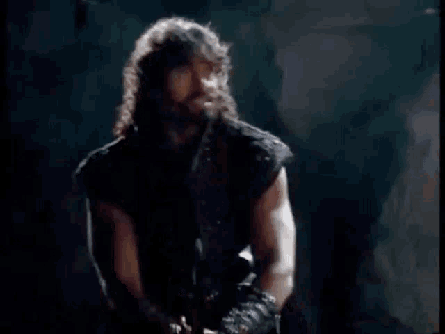 a man with long hair and a beard is standing in a dark room and saying come on .