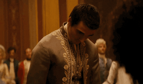 a man in a grey jacket with gold embroidery stands in a room