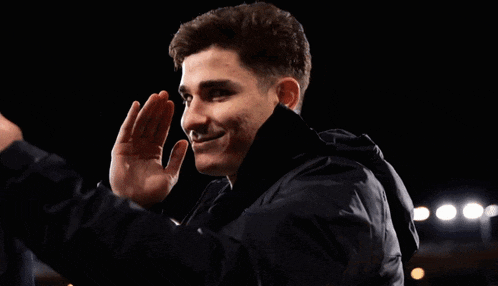 a man in a black jacket is smiling with his hand on his forehead