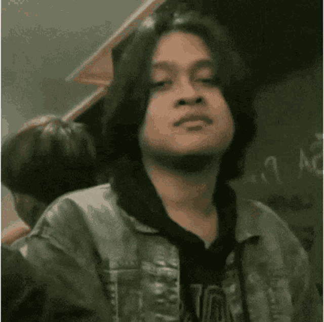 a man with long hair and a beard is standing in front of a blackboard and looking at the camera .