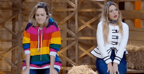two women are sitting next to each other in a barn and one is wearing a rainbow sweater .