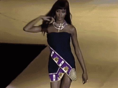 a woman is walking down a runway wearing a black dress and a purple sash