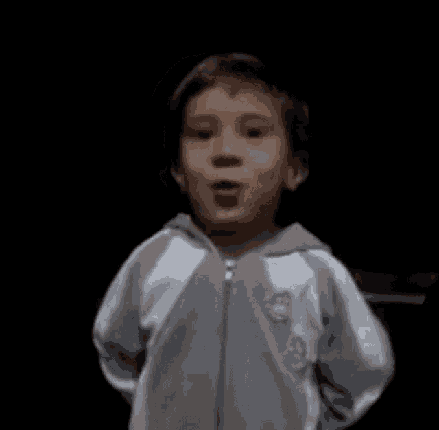 a young boy in a gray and white jacket is making a funny face in a dark room .