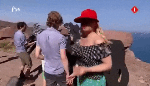 a woman in a red hat is standing on a cliff overlooking the ocean holding hands with a man .