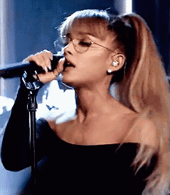 ariana grande is singing into a microphone while wearing glasses and a ponytail .
