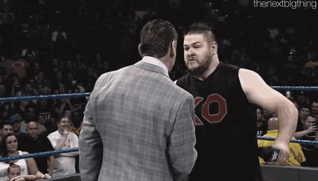 a man in a suit and a man in a black ko shirt are standing in a wrestling ring .
