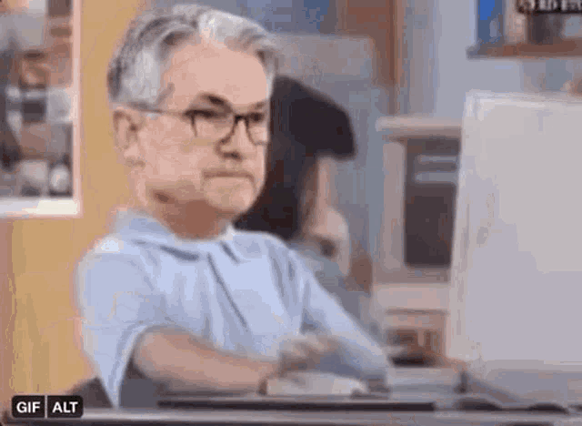 a man wearing glasses is sitting at a desk in front of a computer monitor .
