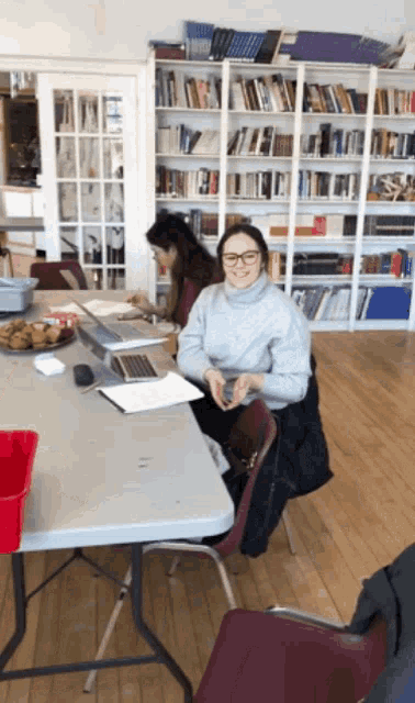 a woman sits at a table in front of a bookshelf