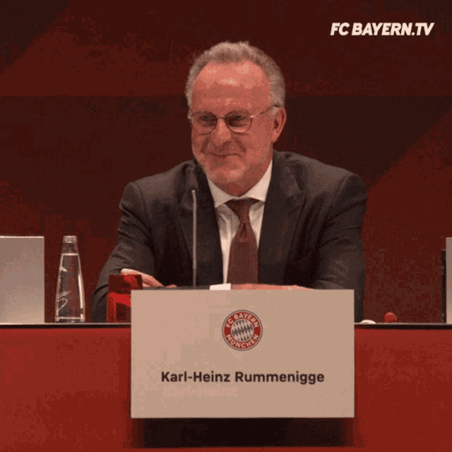 a man in a suit and tie sits at a table with a sign that says karl-heinz rummenigge on it