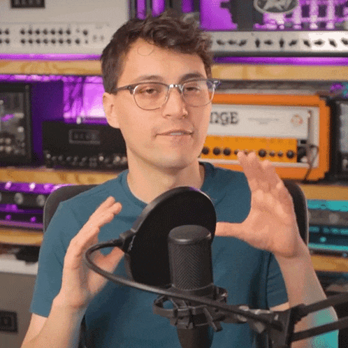 a man wearing glasses and a blue shirt stands in front of a microphone with an orange amplifier in the background