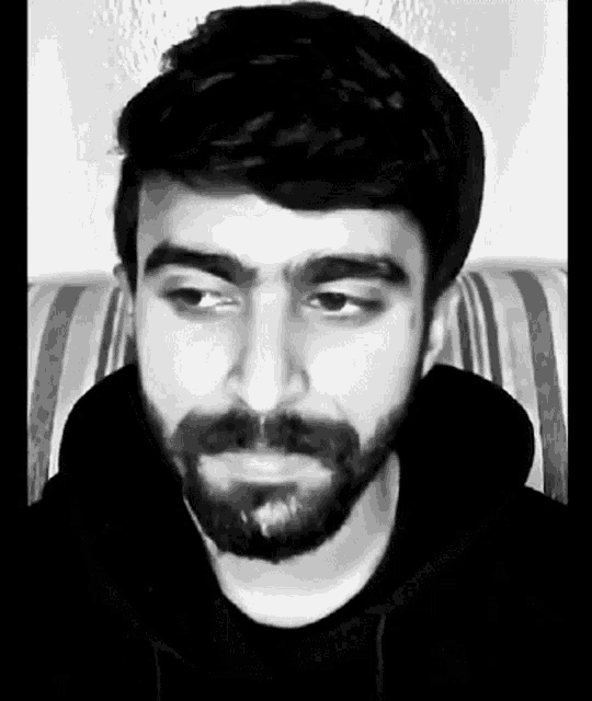 a man with a beard and mustache is sitting on a couch and looking at the camera in a black and white photo .