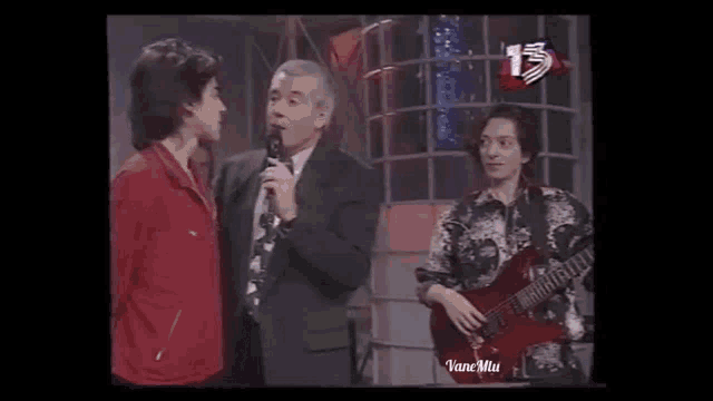 a man in a suit and tie is talking into a microphone while a man playing a guitar stands next to him .