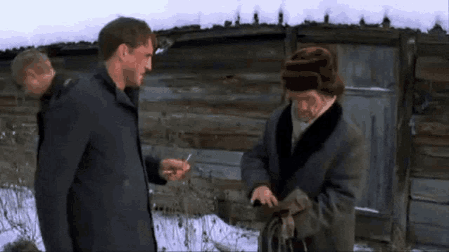 two men are standing in front of a wooden building one is holding a cigarette