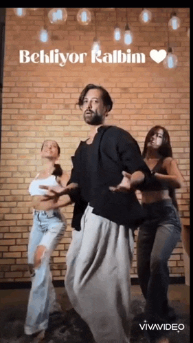 a man and two women are dancing in front of a brick wall with the words behliyor kalbim above them .
