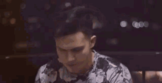 a young man in a floral shirt is sitting in a dark room and making a funny face .